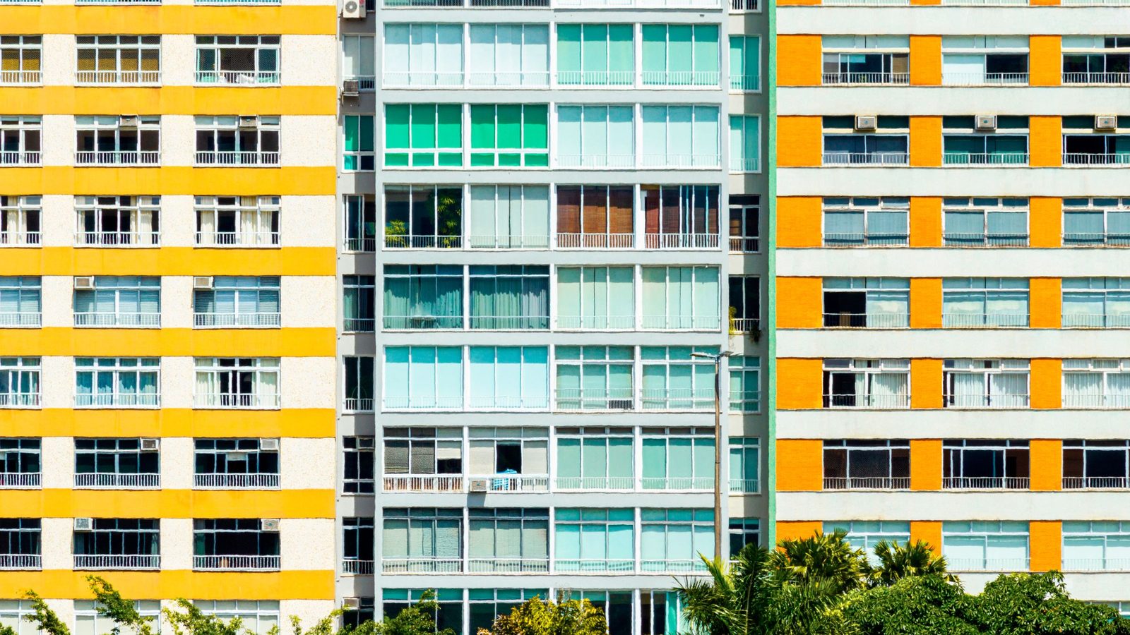 Fachadas de três prédios residenciais modernistas no Rio de Janeiro, em faixas amarelas, verde-água e laranja, com janelas em banda e palmeiras e árvores tropicais junto à base. Imagem ilustrativa, não é um imóvel analisado pela Rivesta.