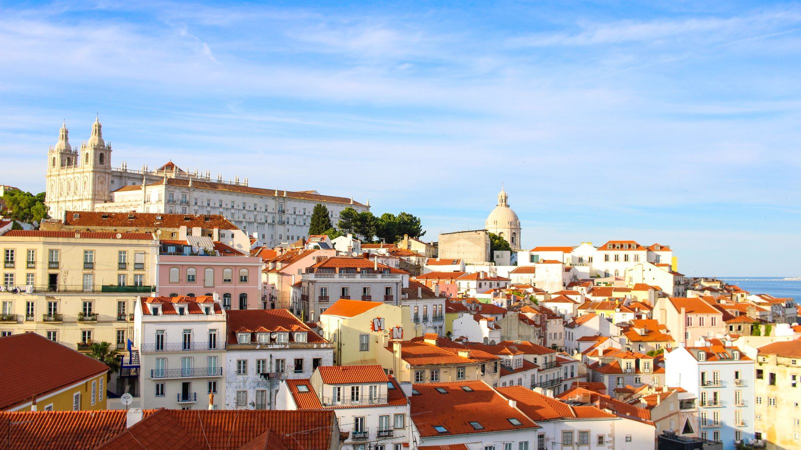 Vista sobre os telhados e prédios de habitação de Alfama, em Lisboa, com São Vicente de Fora e o Panteão ao fundo, sob céu limpo. Imagem ilustrativa, não é um imóvel analisado pela Rivesta.